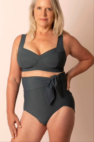 Larger mature woman wearing a dark blue-gray, high-waist two-piece bikini set on a beige background