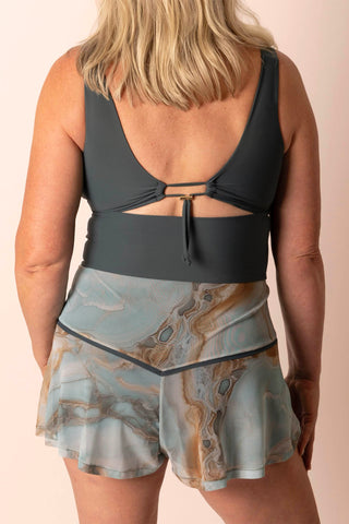 Back view of a larger mature woman wearing a dark blue-gray swim top with matching light teal and tan marble printed swim skort skirt.