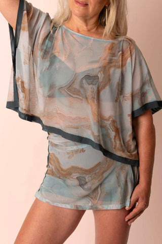 Larger mature woman wearing a flowy sheer mesh rectangle top in a light teal and tan marble print with dark blue-gray contrast. Top is paired with dark blue grey swim top and swim skort skirt with matching print. 